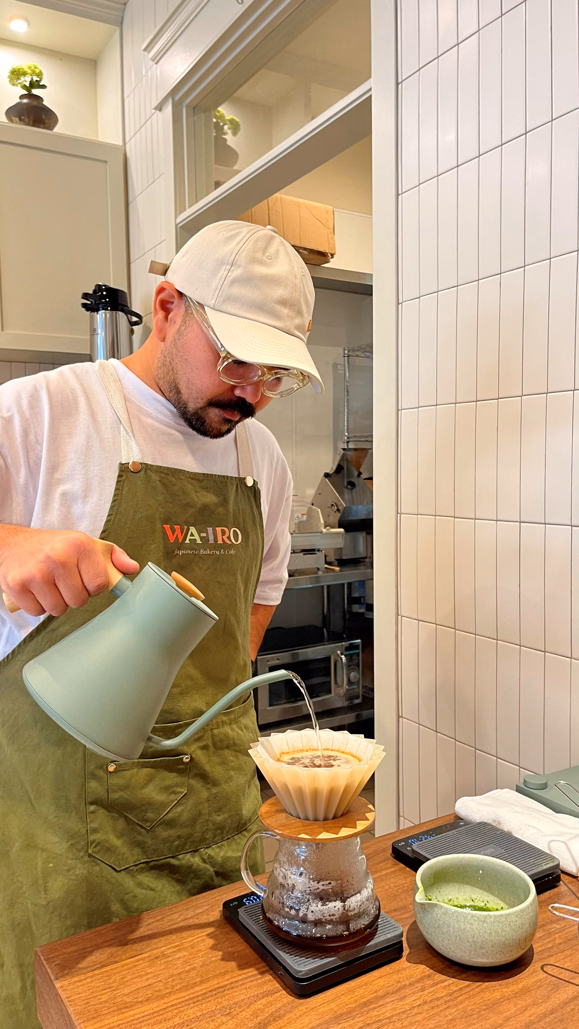 A staff member finishing a hand-whisked matcha pour-over, kettle in hand beside the ceramic bowl and dripper.
