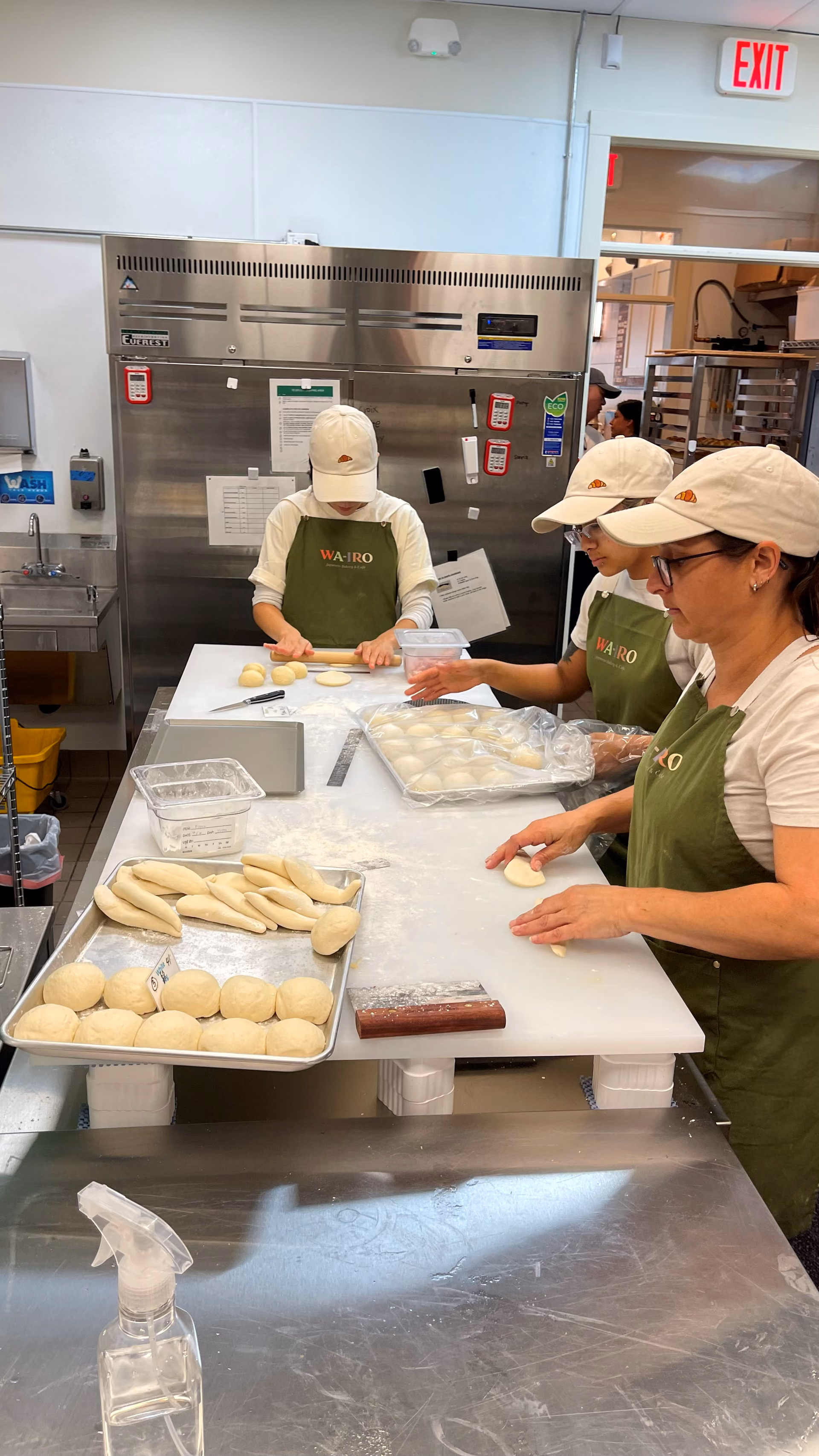 Three staff members shaping bread dough by hand at a stainless steel prep table, trays of proofed rolls beside them.