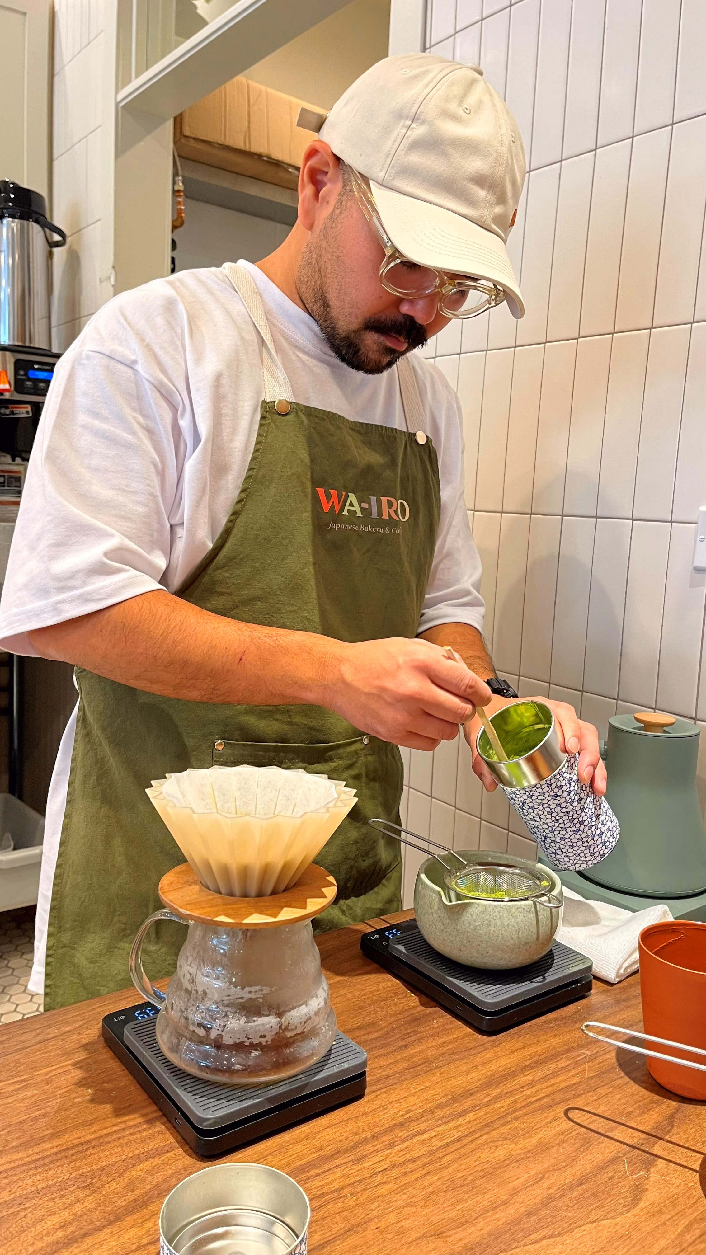 A staff member in a WA-IRO apron pouring hot water over a matcha pour-over setup, whisked matcha visible in a ceramic bowl.