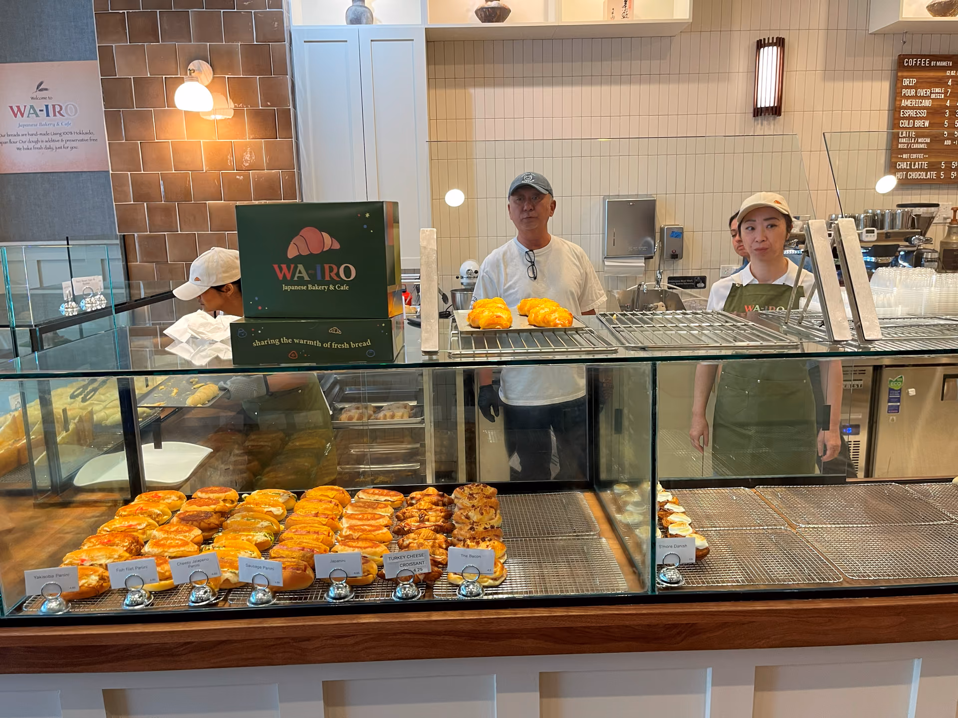 Two staff members behind a glass pastry case filled with golden croissants and pastries, a WA-IRO branded box on the counter.